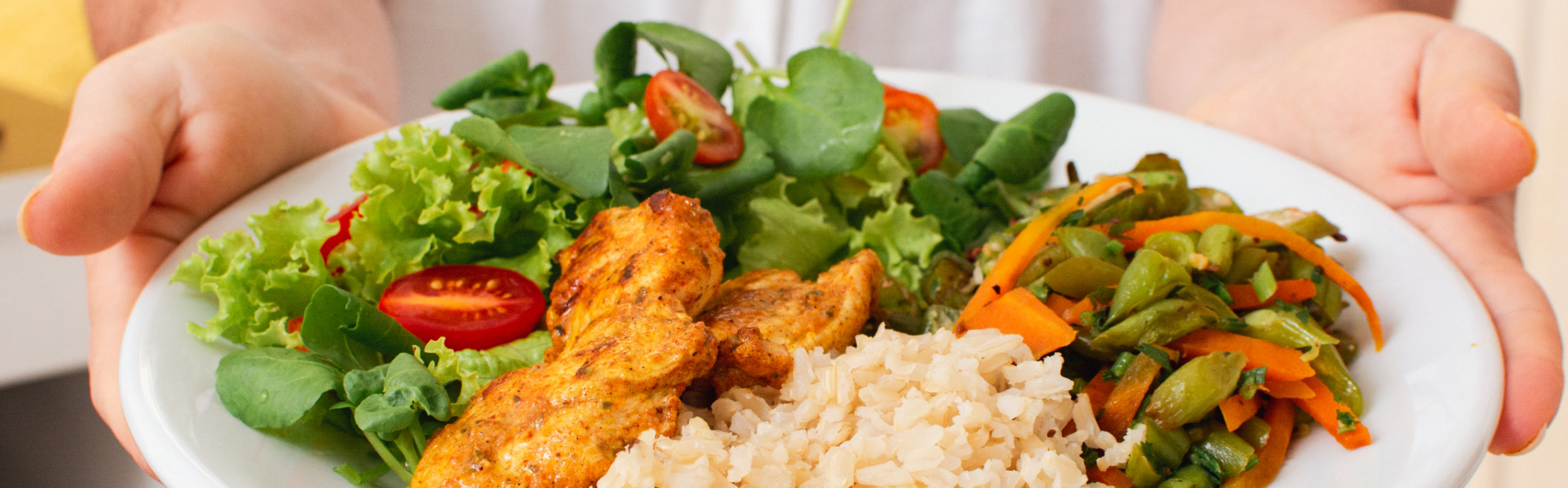 A person holding a plate with a meal consisting of seasoned chicken, brown rice, mixed salad greens, cherry tomatoes, and sautéed vegetables.