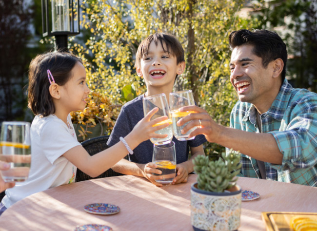 A family of four smiling while drinking fruit-infused water at a table in a sunny backyard.