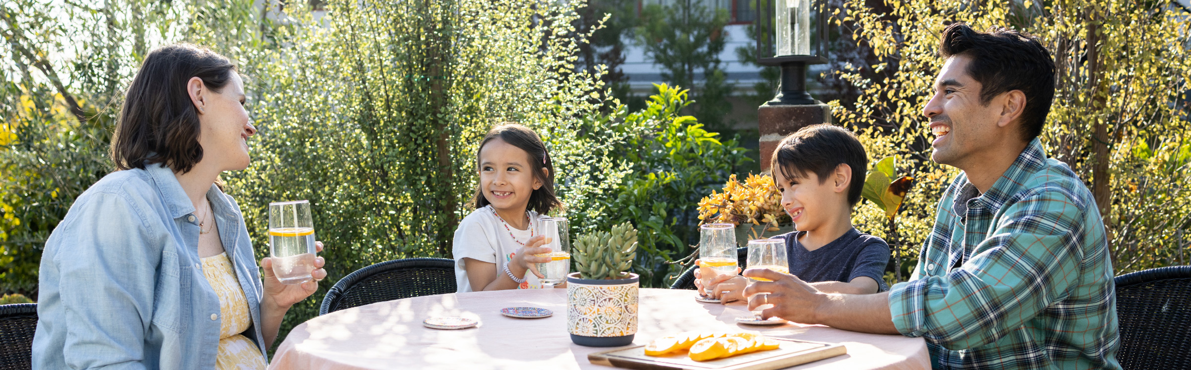 Una familia de cuatro a la mesa sonriendo mientras beben agua con frutas en un jardín soleado.